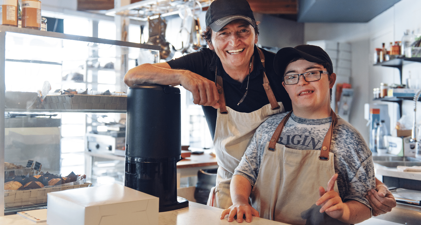 accessible workplaces: Two cafe workers stand behind the counter, one is a man with down syndrome.