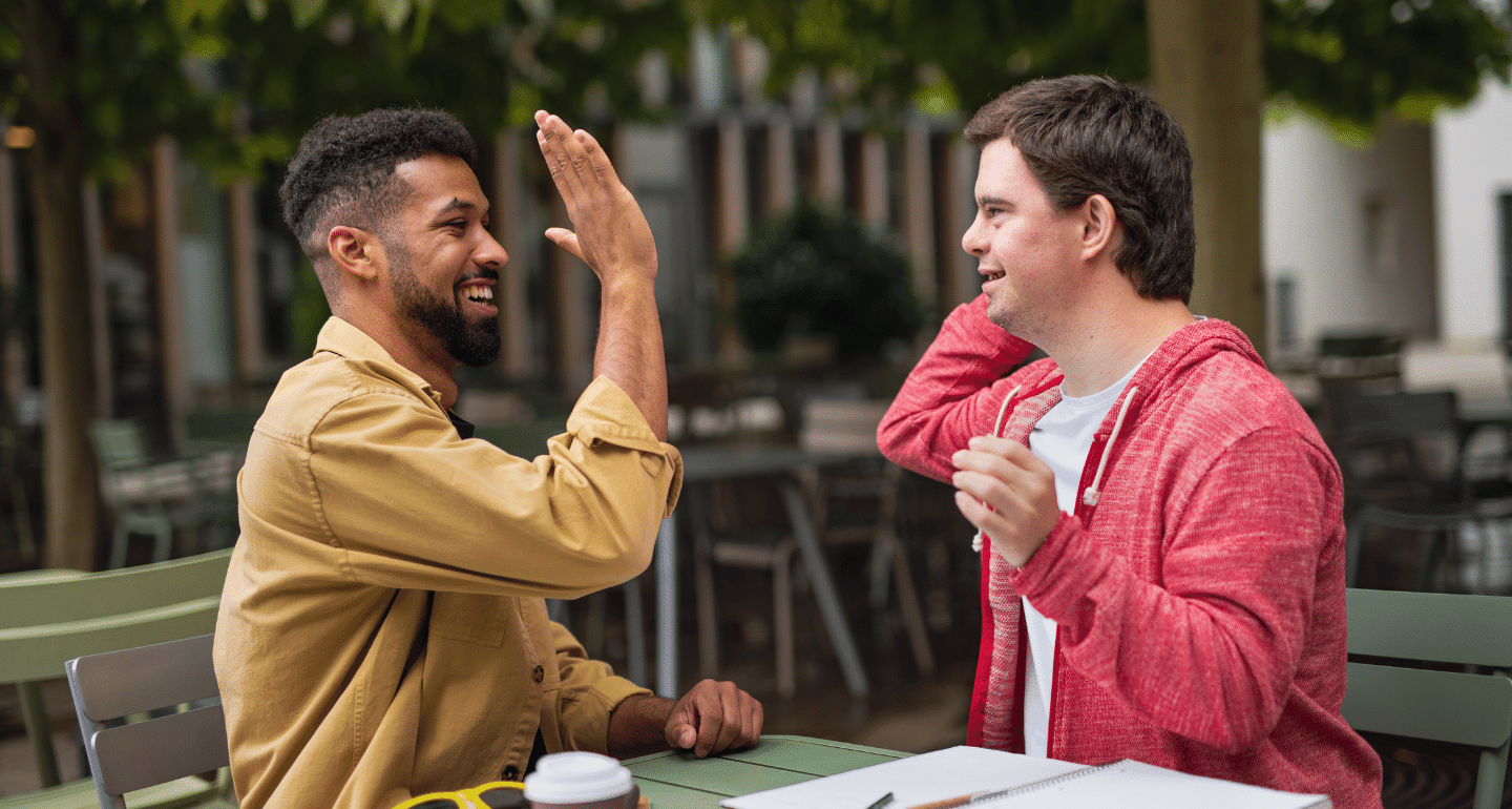 Supported Decision-Making: Male support worker high fives man with a disability at a cafe.