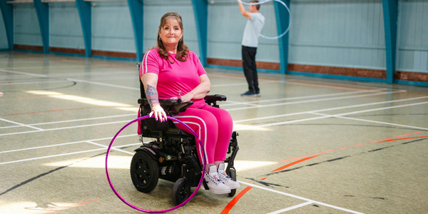 Woman in a hot pink outfit using a motorised wheelchair and hula hoop in a gym, showing NDIS assistive technology supporting active, independent living