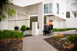 Short-Term Accommodation (STA): Man in an electric wheelchair on a path in front of a house.