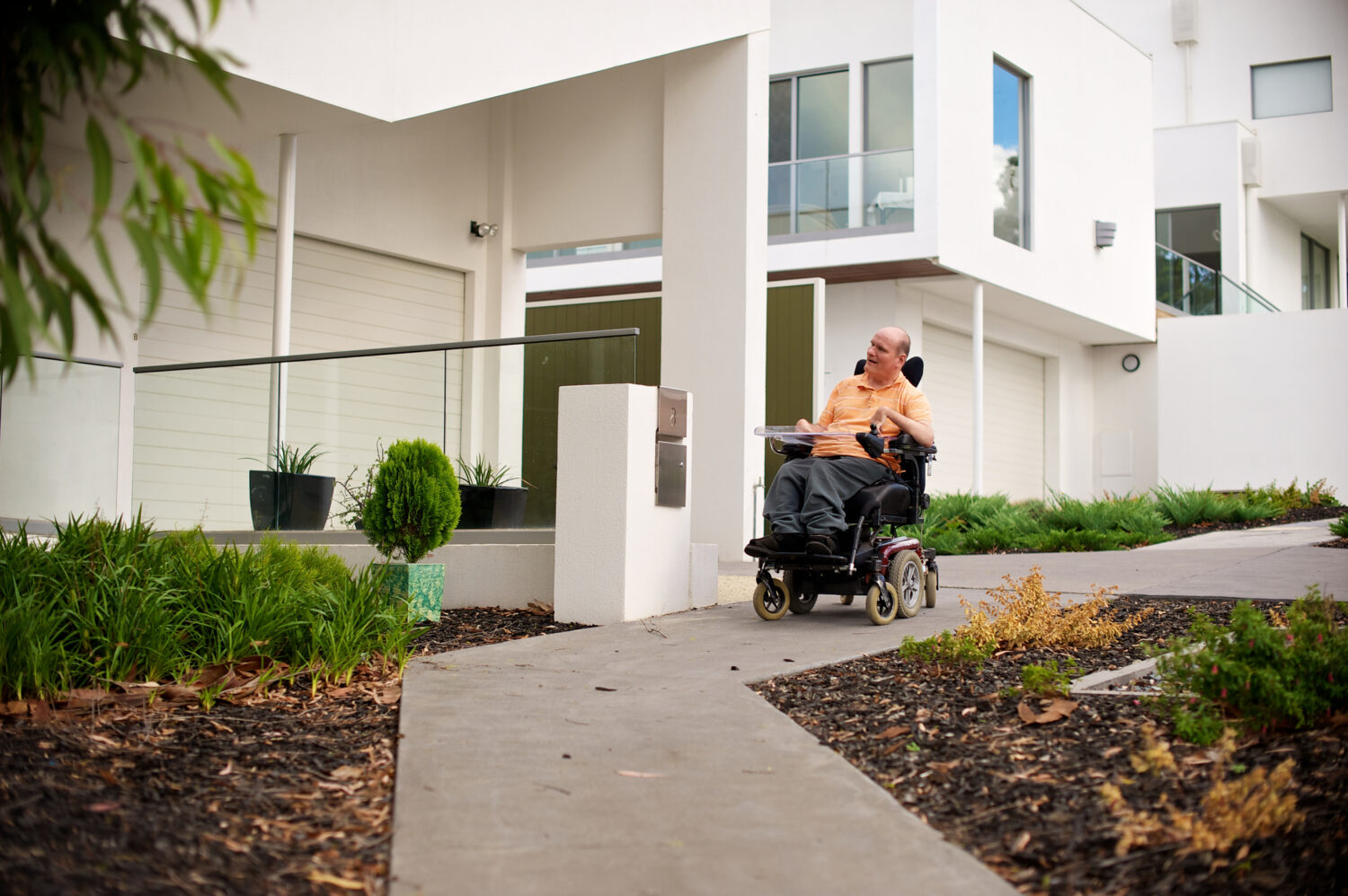 Short-Term Accommodation (STA): Man in an electric wheelchair on a path in front of a house.