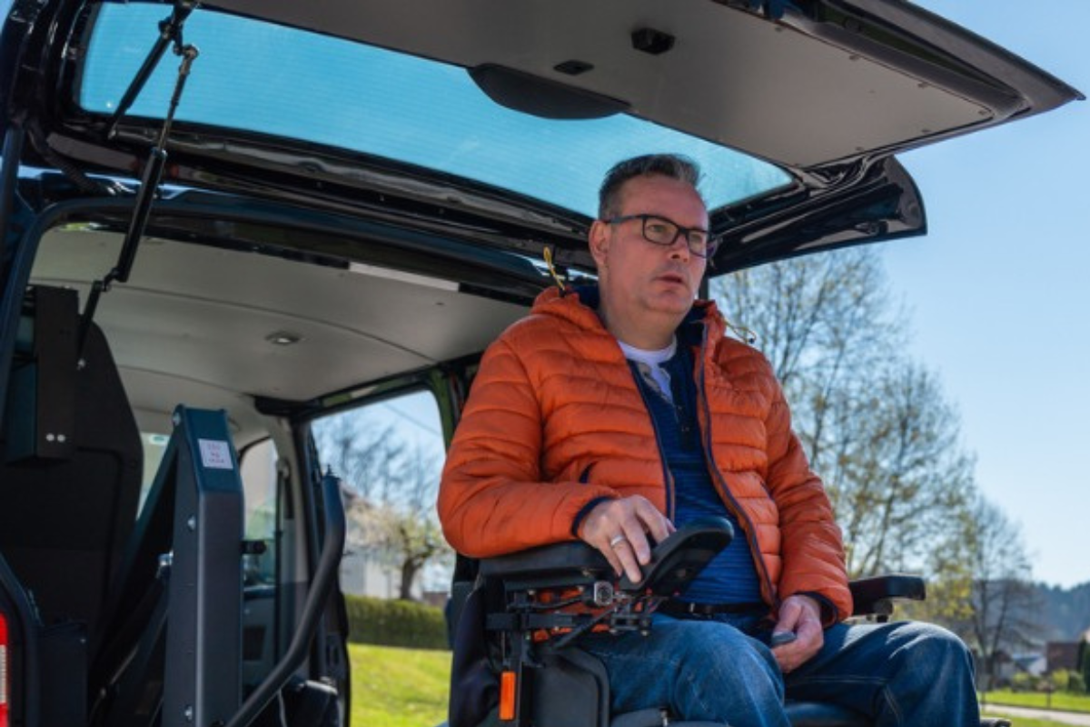 Man using a motorised wheelchair exits a wheelchair-accessible vehicle with support from a complex disability care service.