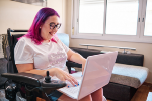 How NDIS Funding Works: Woman with pink hair using an electric wheelchair, logging into her NDIS portal on a pink laptop at home