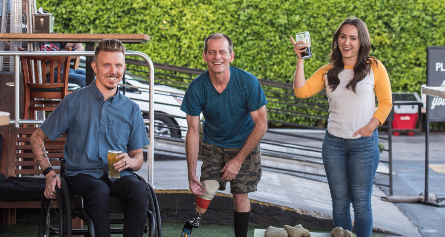 Social and Community Participation: Man with a prosthetic leg and a male wheelchair user playing cornhole with a female friend, enjoying a fun outdoor activity together.