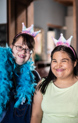 day programs: Two women with intellectual disabilities smiling in crowns and feather boas at an NDIS day program.