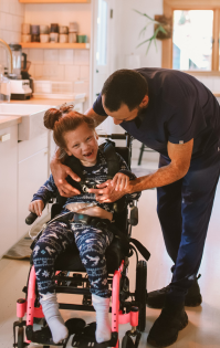 Carer and Family services referrals: A father lovingly cares for his young daughter in a wheelchair as they spend time together in the kitchen, highlighting the importance of family support and caregiving.